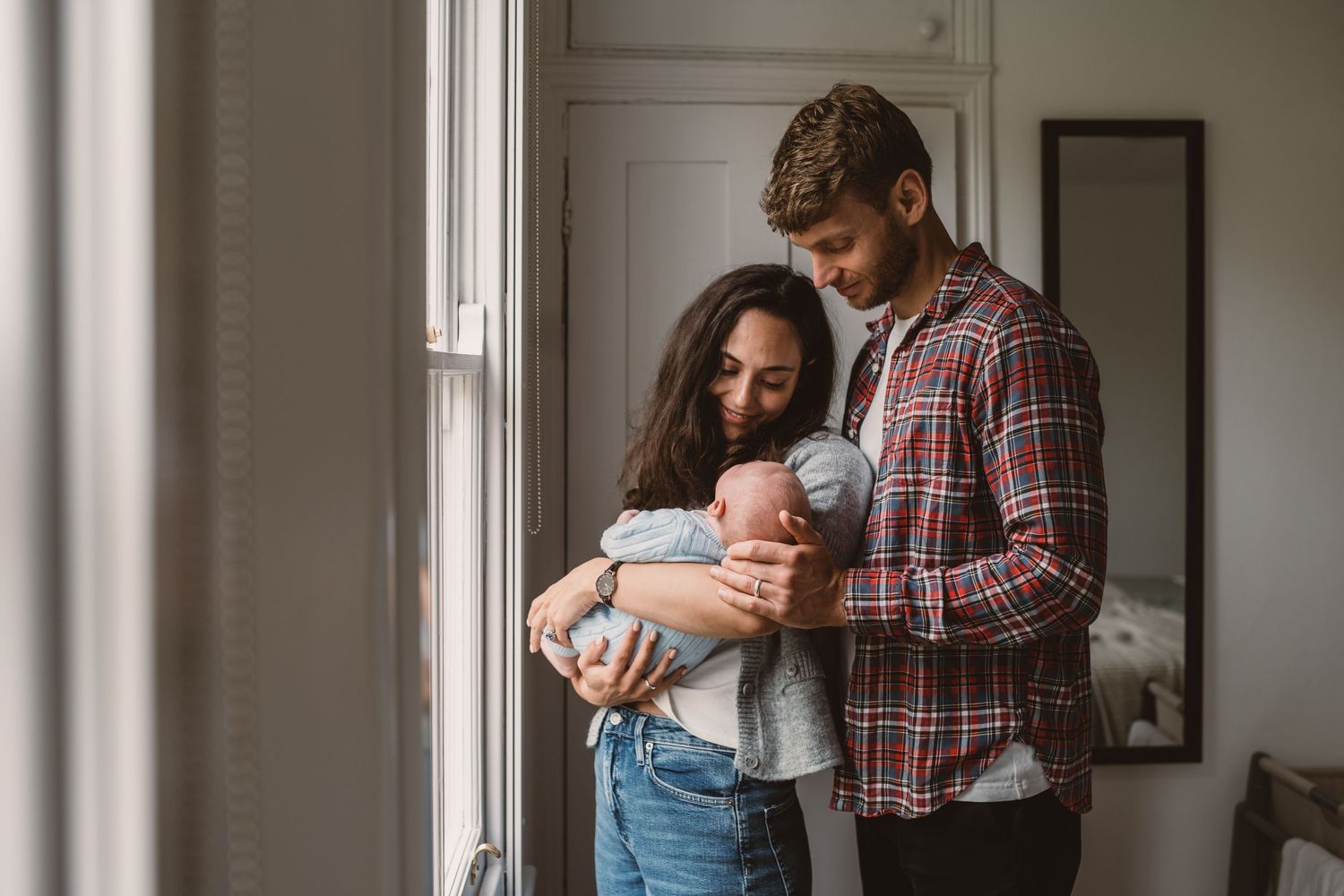 Young couple standing by window holding their newborn baby, sharing a tender family moment at home.