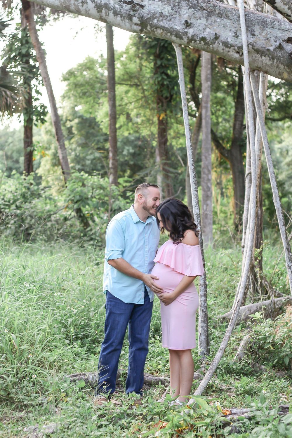 Couple sharing a tender moment outdoors, pregnant woman in pink dress with partner in blue shirt, surrounded by lush greenery at Riverbend.