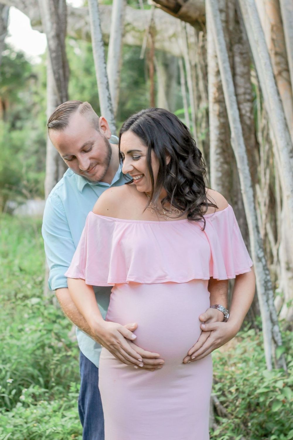 Couple in maternity photo shoot outdoors, woman in pink off-shoulder gown, man in blue shirt, both cradling baby bump at Riverbend Park.