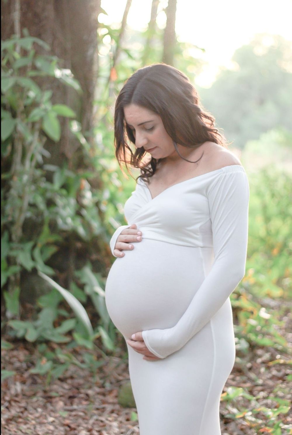 Pregnant woman in white off-shoulder gown cradling baby bump outdoors among lush greenery in soft natural light at Riverbend Park Jupiter.