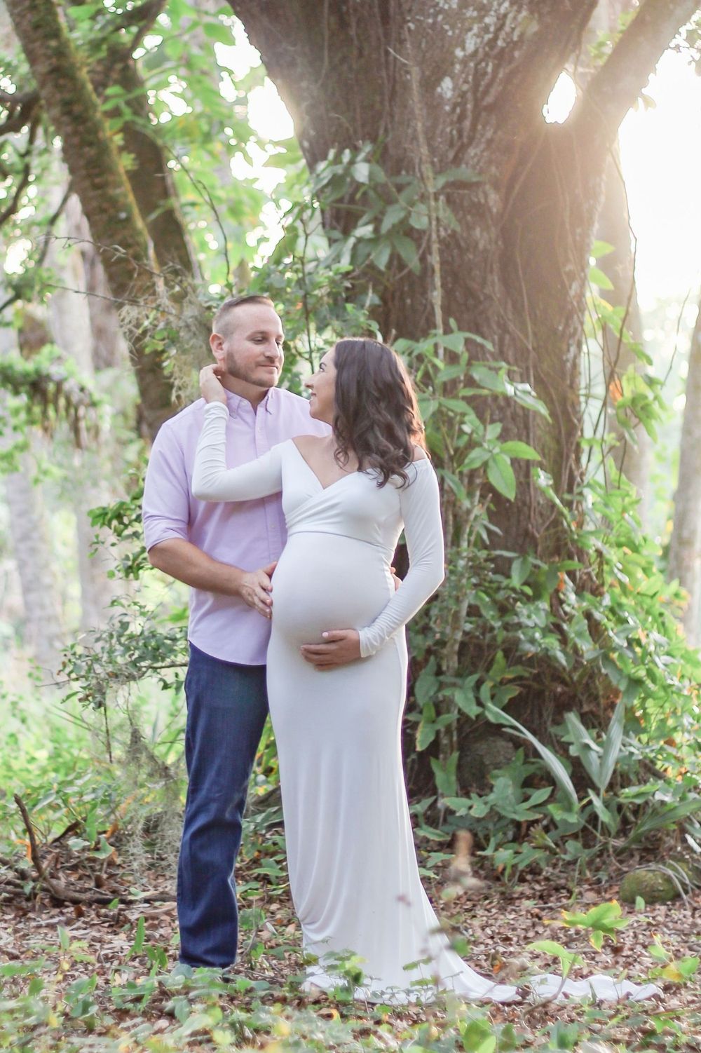 Couple poses outdoors for maternity photo shoot, pregnant woman in white gown cradled by partner near sunlit trees at Riverbend Park Jupiter