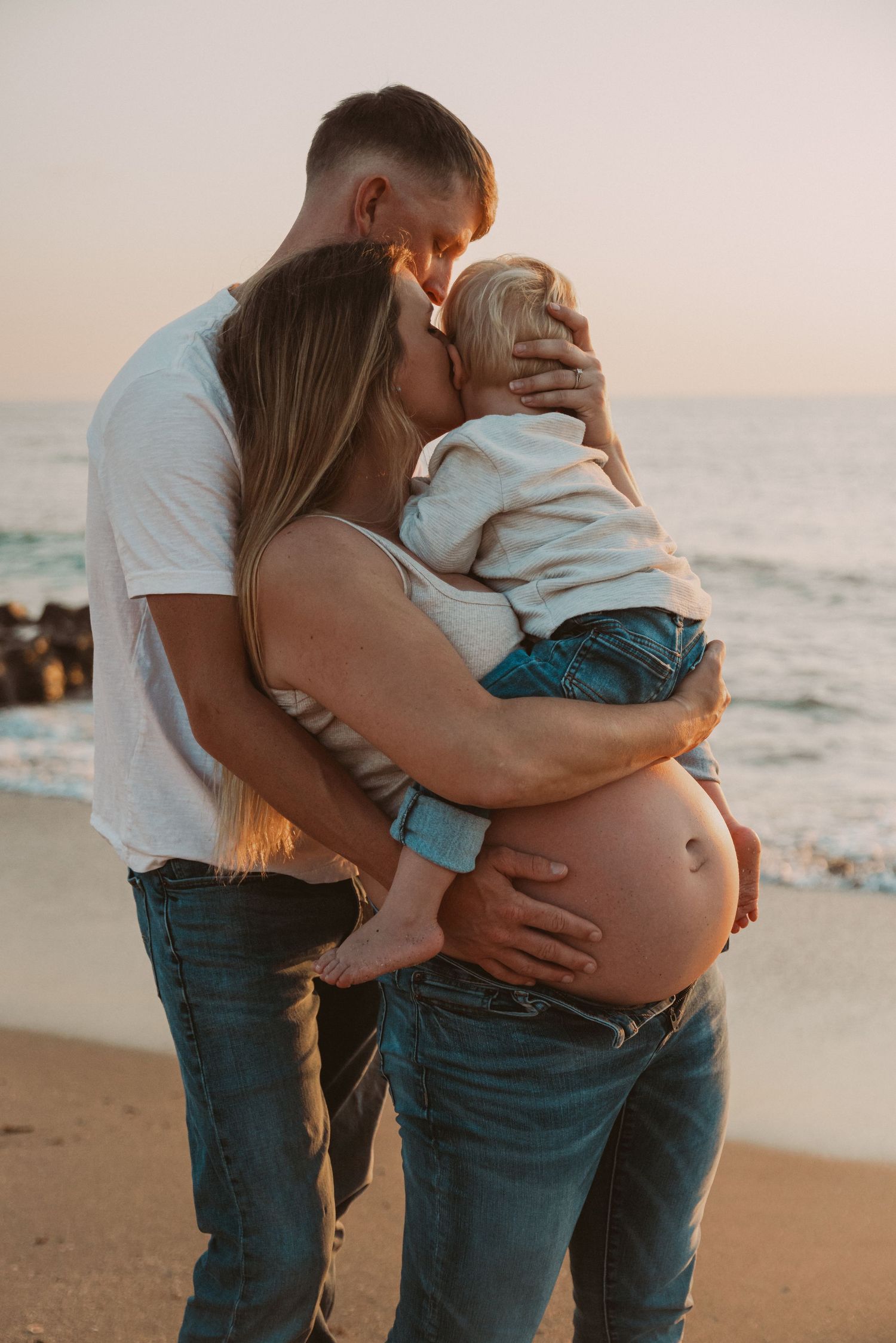 Intimate family moment on beach at sunset shows pregnant woman's belly while holding toddler with partner.