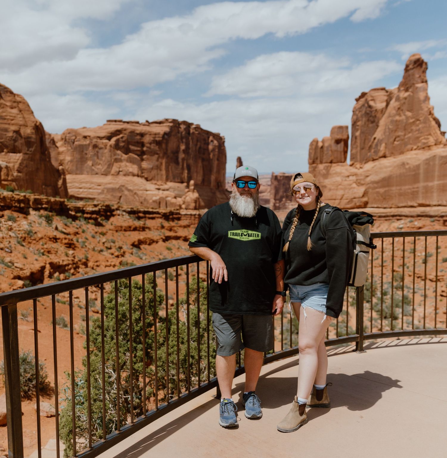 Sydney and her dad pose along a railing at Arches National Park with towering red rock formations in the background