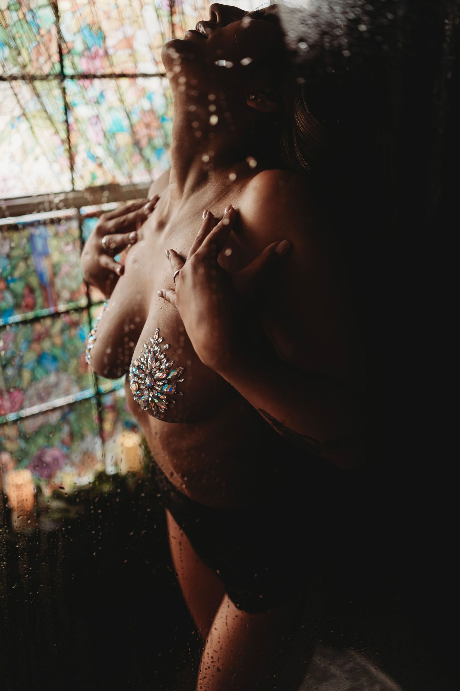 A woman sensually posing in a shower with stained glass in the background. Her chest is adorned with delicate body jewelry, and steam softly