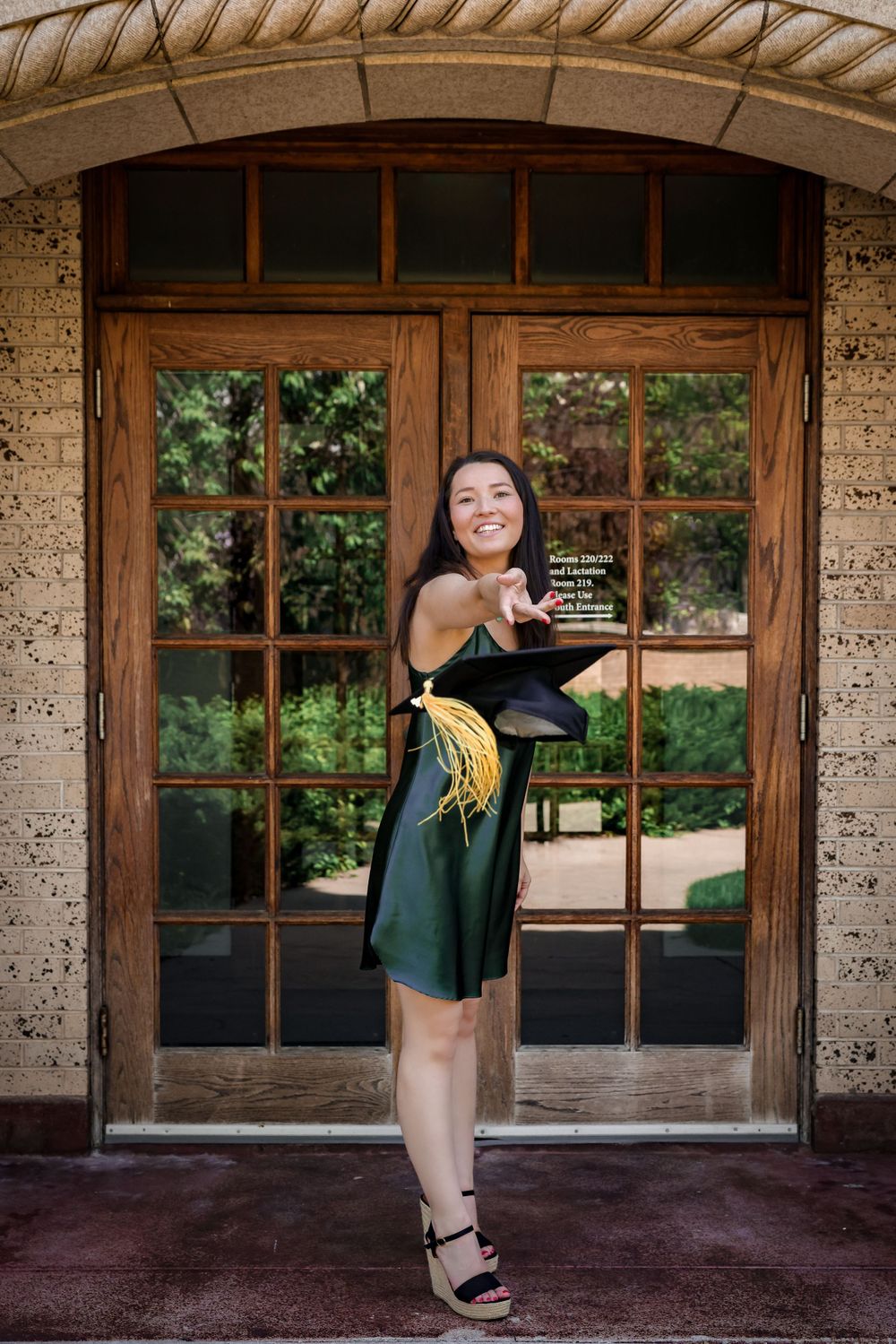 Person in green dress poses with graduation cap in front of ornate wooden door.