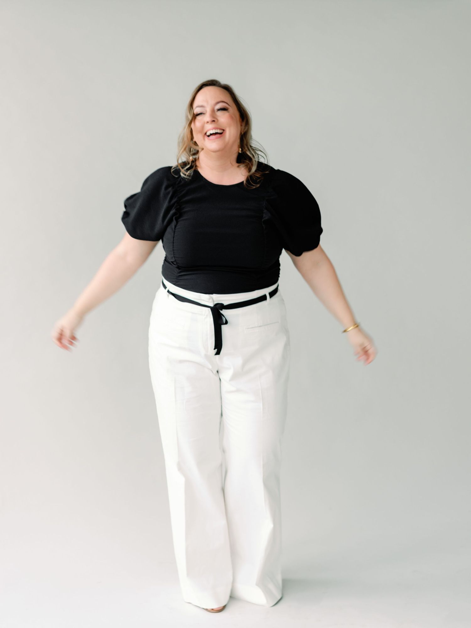 A person in a black puff sleeve top and white wide-leg pants poses joyfully against a light gray background.