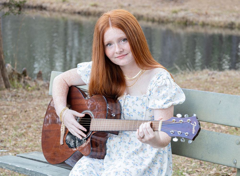 A musician in a white floral dress plays acoustic guitar while sitting on a bench near a lake.
