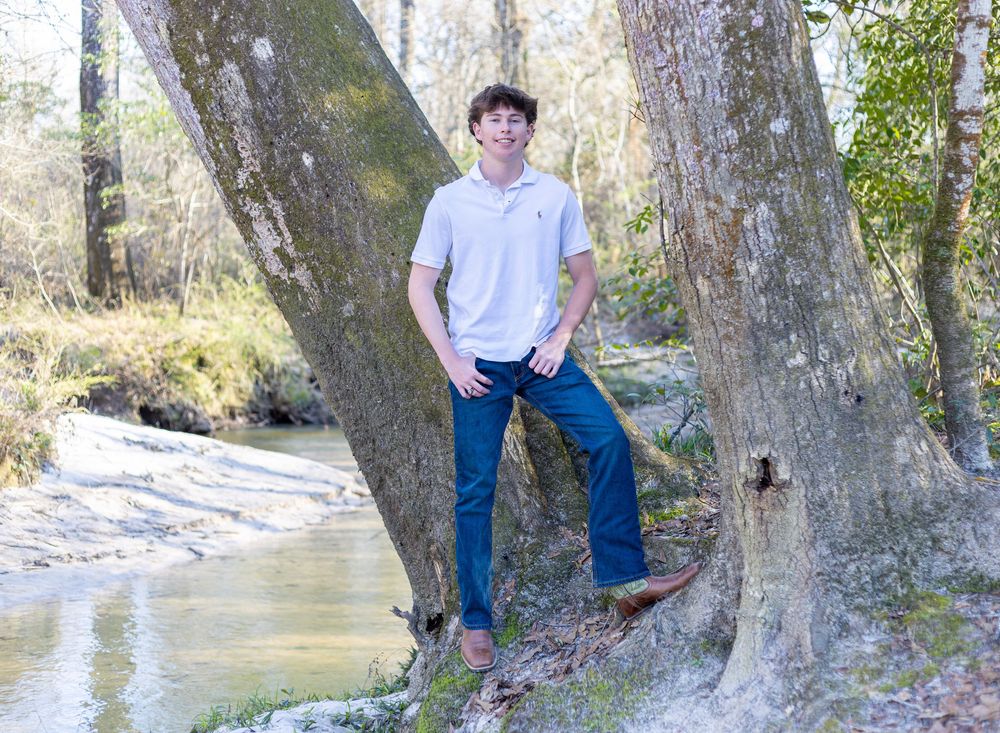 A casual outdoor portrait by a creek with someone leaning against a large tree trunk in jeans and white shirt.
