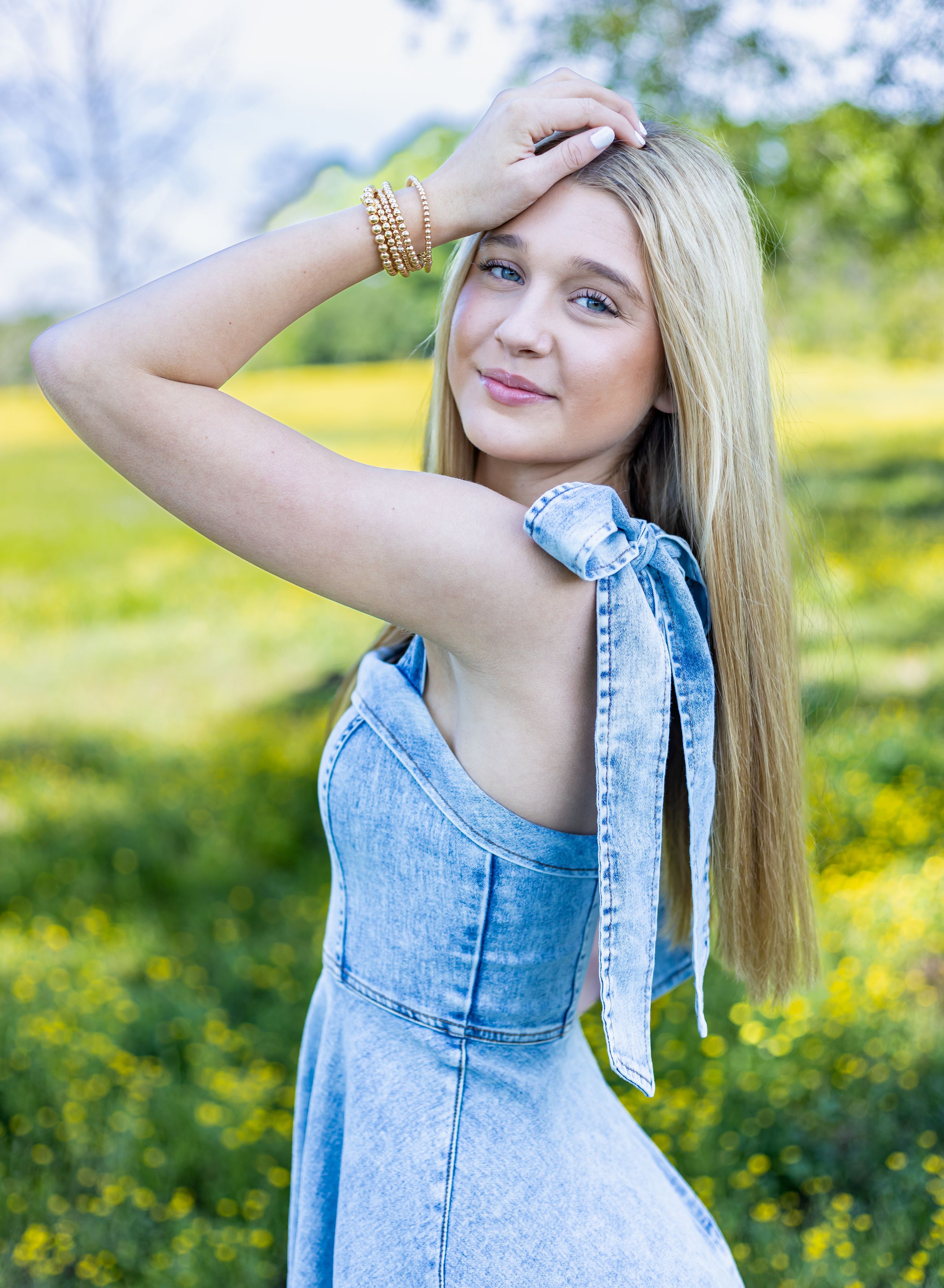 A person in a denim dress poses outdoors in a sunny meadow with yellow wildflowers and greenery in the background.
