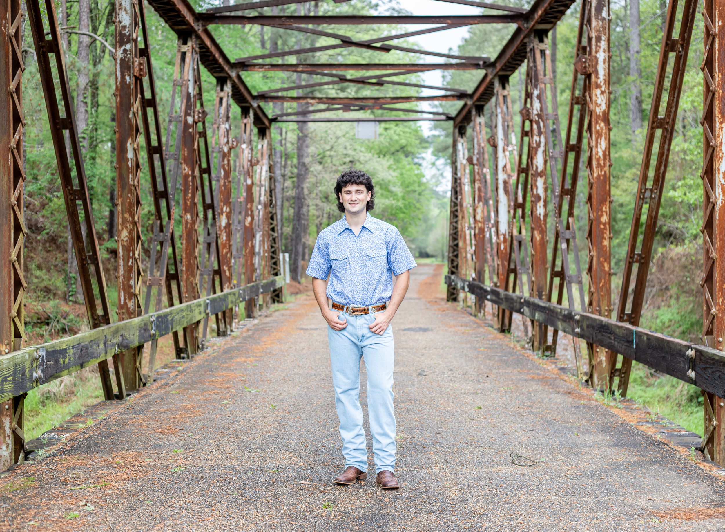 A rustic iron bridge spans a wooded path with someone standing in the center wearing jeans and a blue shirt.