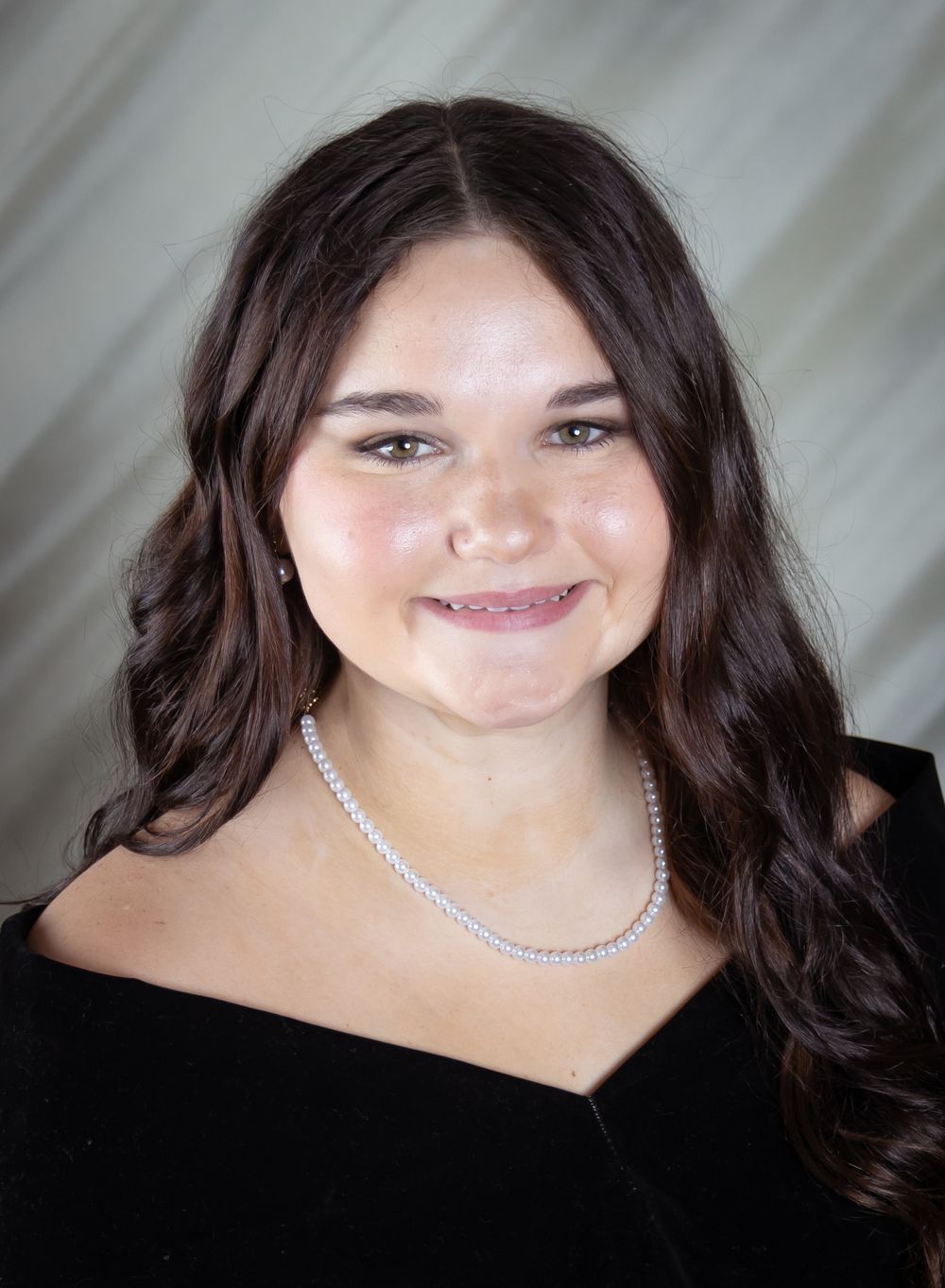Young woman with dark wavy hair smiling, wearing a black top and pearl necklace, formal portrait photo.