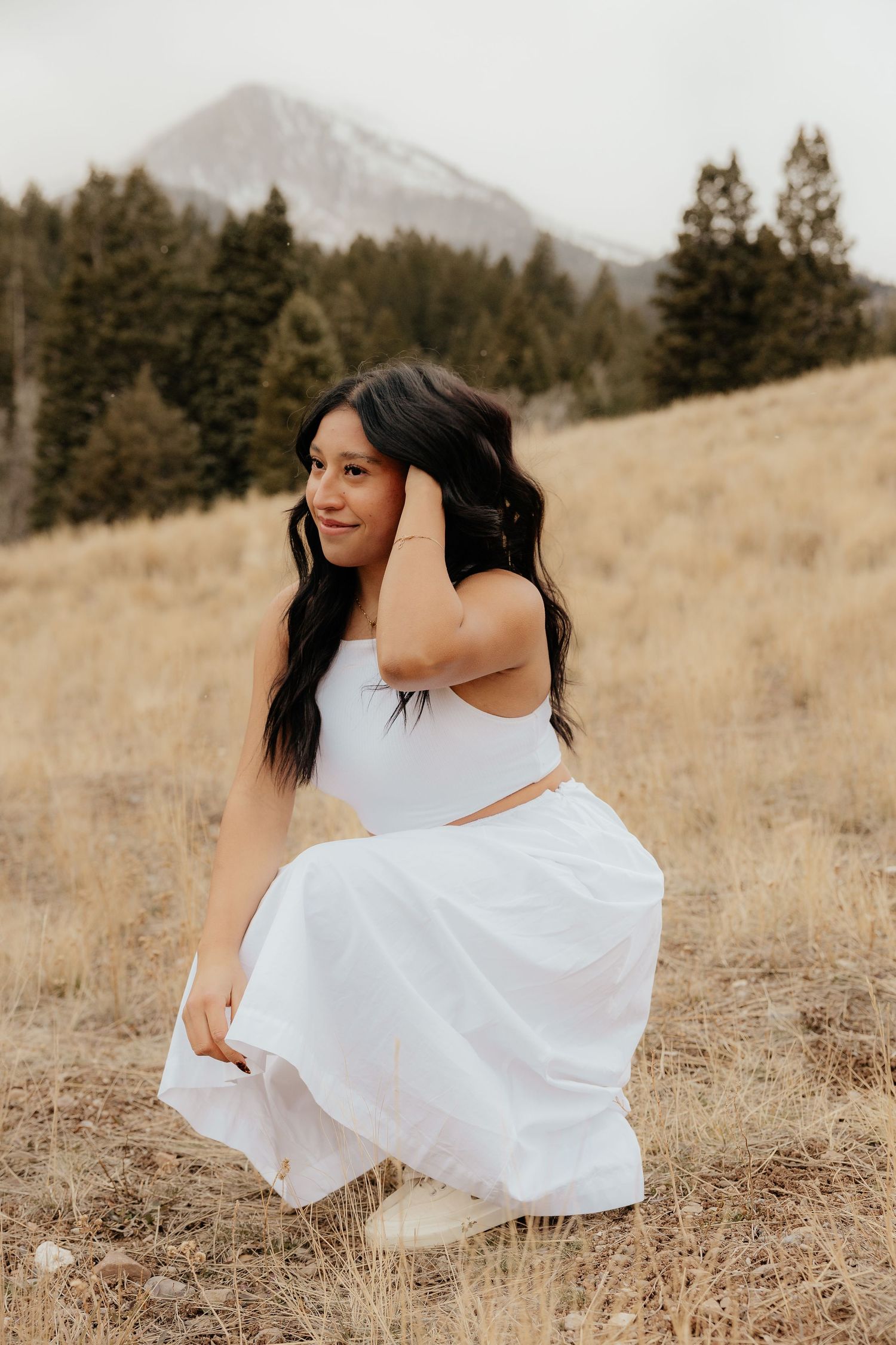 Young woman in white crop top and skirt posing in a golden field with mountain and pine trees behind.