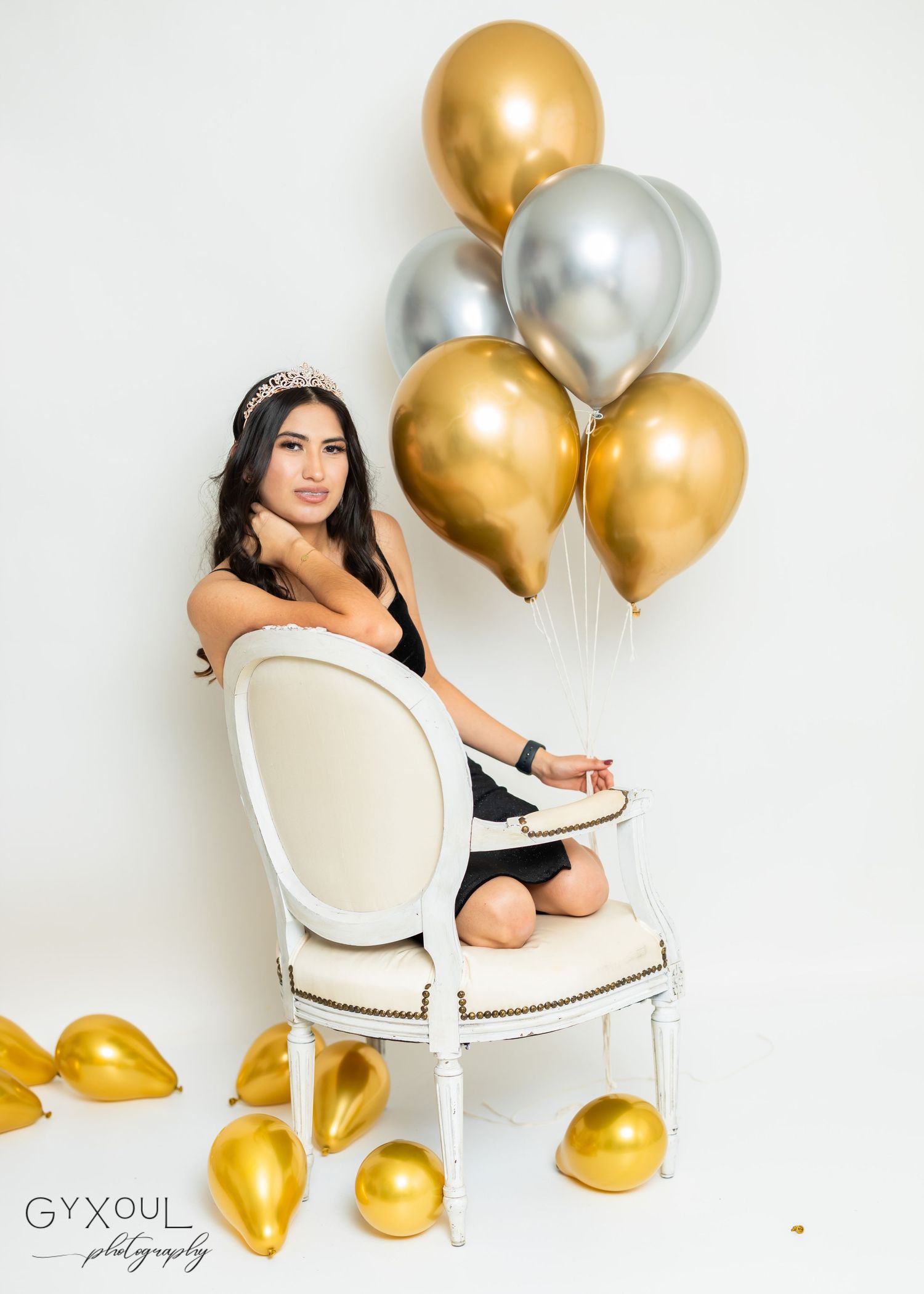 Elegant pose on white chair with gold and silver balloons in background.
