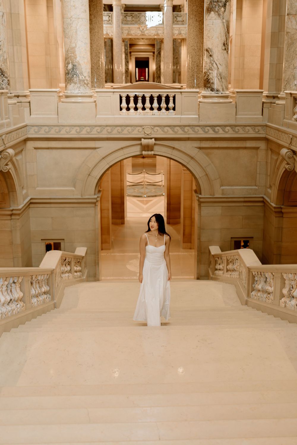 A person in an elegant white dress stands centered in a grand classical architectural setting.