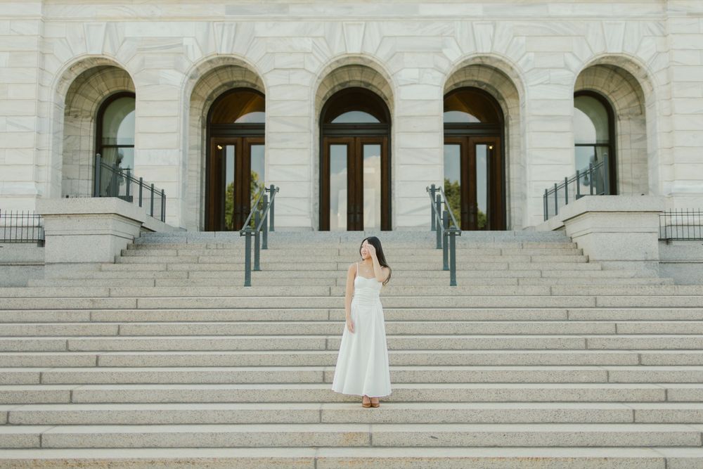 Person in flowing white dress standing on wide stone steps of a neoclassical building.