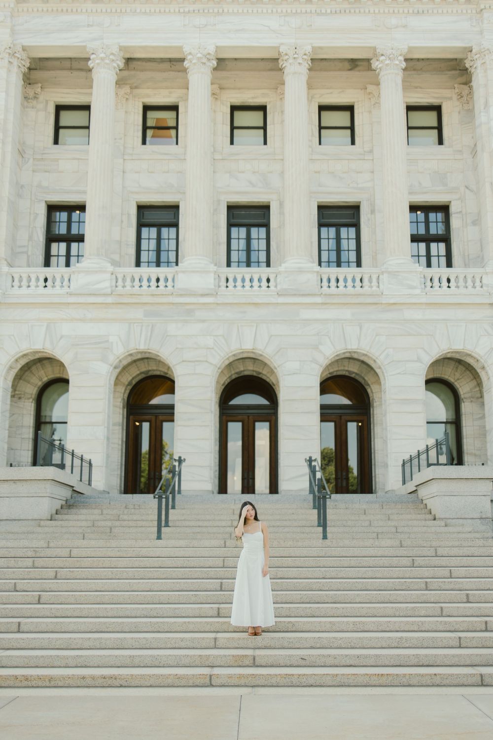 White dress contrasts against classical architecture with columns and grand staircase.
