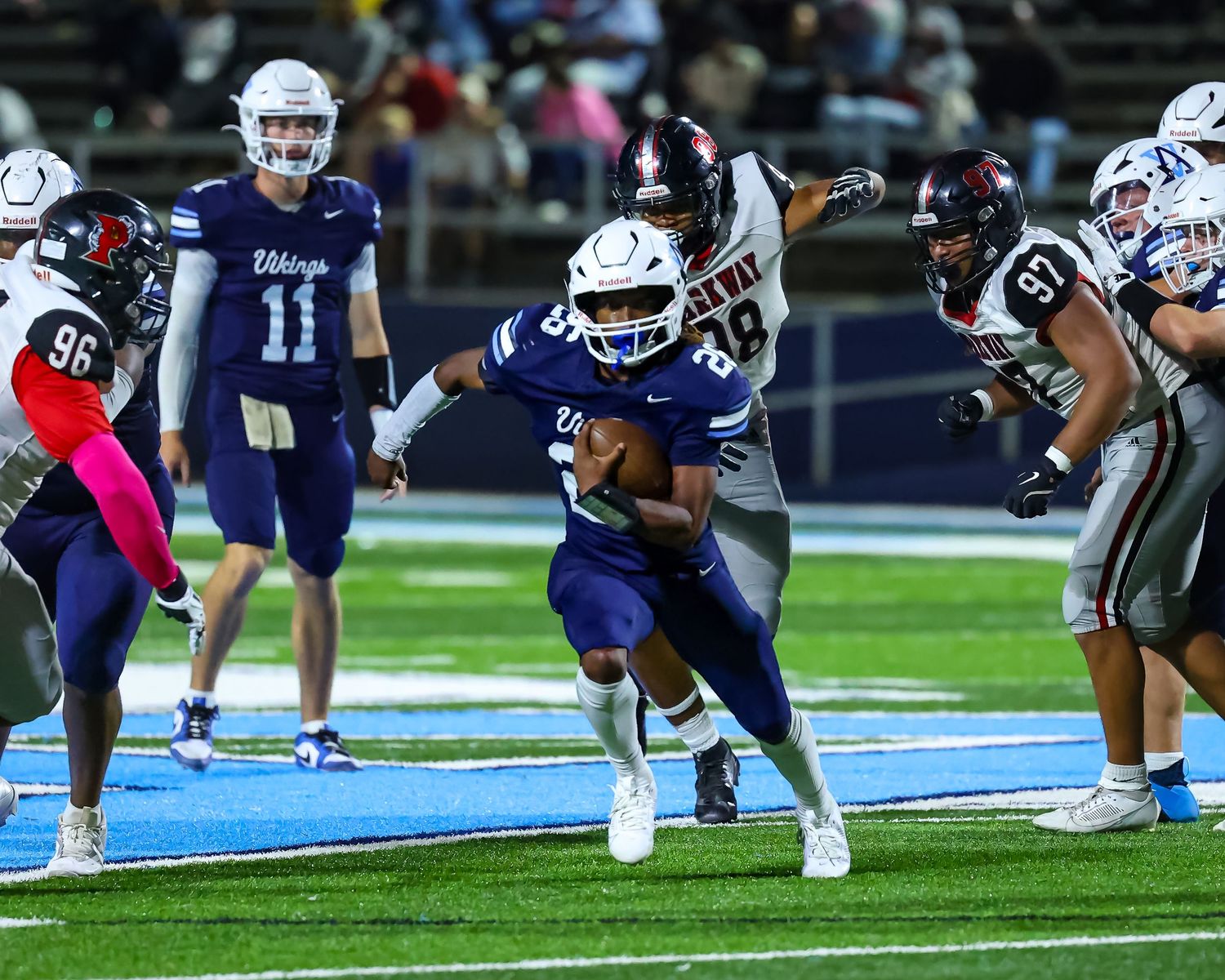 High school football action shows a running back in blue uniform breaking through tacklers on blue turf during night game.