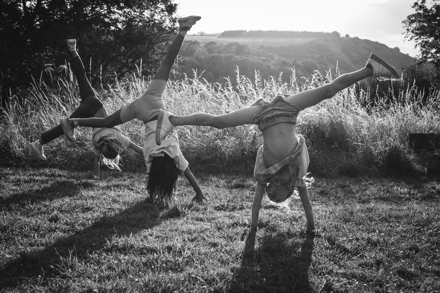 Three children doing handstands on grass with mountains in background, captured in black and white.