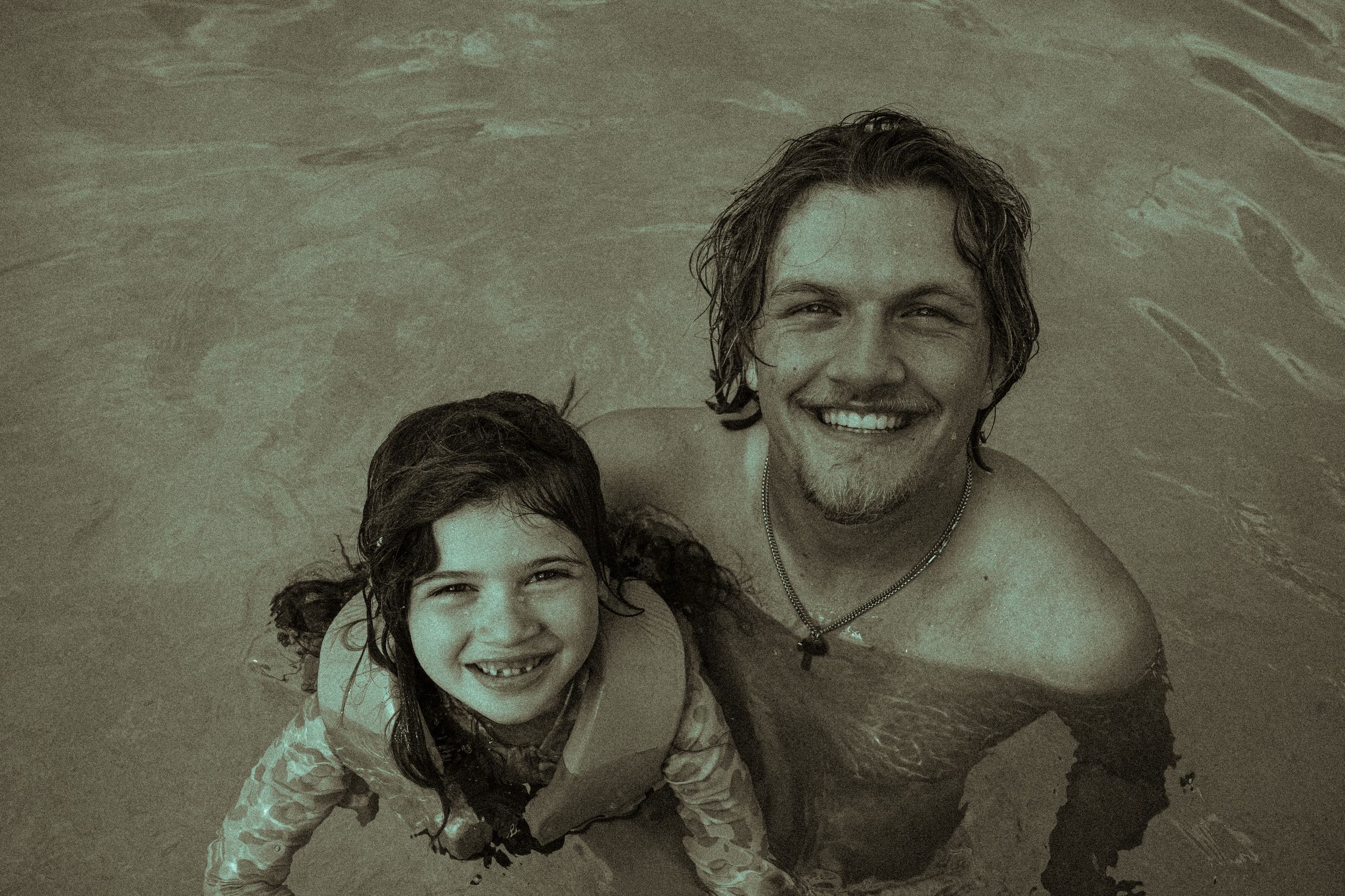 A smiling man and young girl pose together in a swimming pool in a vintage-toned photo.