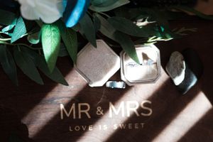 Wedding rings and 'MR & MRS' text display with green foliage in moody lighting for romantic wedding detail shot.