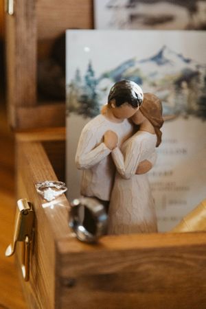 A Willow Tree figurine of two figures embracing stands on a wooden surface with a mountain painting in the background.