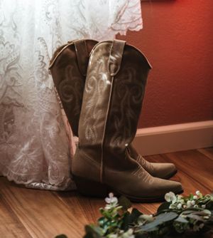 A brown leather cowboy boot sits next to white lace fabric and floral decorations on a hardwood floor.