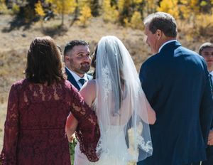 An outdoor wedding ceremony takes place against a backdrop of autumn foliage in the mountains.