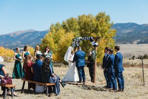 Outdoor mountain wedding ceremony with guests seated on benches under autumn trees.