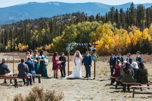 Mountain wedding ceremony with guests gathered under decorated arch amid fall foliage.