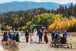 An outdoor mountain wedding ceremony with guests gathered in a field surrounded by autumn foliage and evergreen trees.