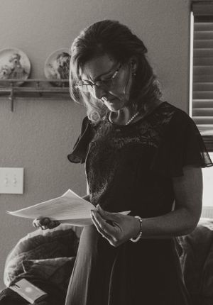A black and white photograph of someone in a dark dress reading papers while standing in a room with decorative plates on a shelf.