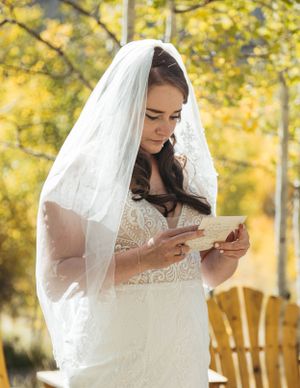 A bride in a white lace dress and veil reads a letter against a backdrop of autumn foliage.