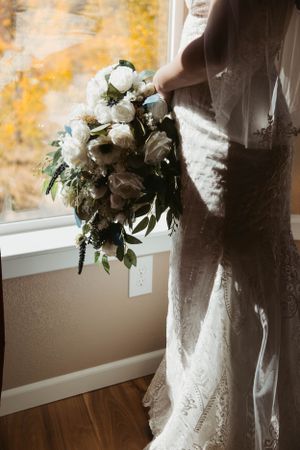 A white bridal bouquet with roses and greenery is held against a lace wedding dress near a window.