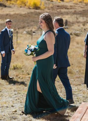 A bridesmaid in an elegant emerald green dress walks outdoors holding a bouquet against a golden autumn hillside.