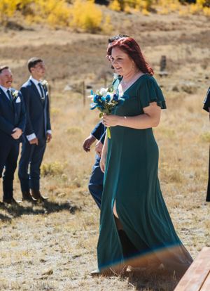 A bridesmaid in an emerald green dress walks across a field with autumn foliage in the background.
