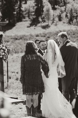 A wedding ceremony takes place outdoors with a bride in a white dress and veil surrounded by guests.