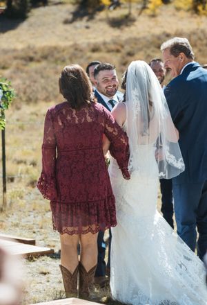 An outdoor wedding ceremony with a bride in white lace and attendees in burgundy and blue against a natural backdrop.