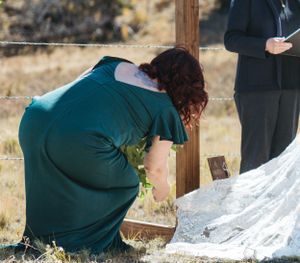 Person in green dress bends down near wooden post during outdoor ceremony.