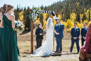A mountain wedding ceremony takes place outdoors with evergreen trees and golden aspens in the background.
