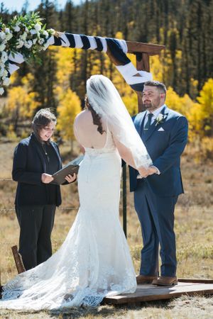 A wedding ceremony takes place outdoors with fall foliage in the background beneath a rustic wooden arch.
