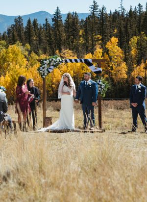 An outdoor wedding ceremony takes place in a mountain meadow with golden autumn trees and evergreens in the background.