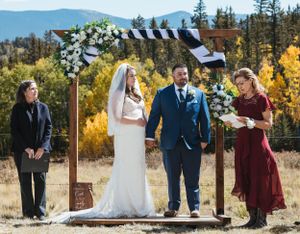 A mountain wedding ceremony under a floral arch with burgundy and white decor against an autumn landscape.