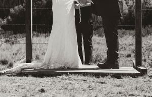 Black and white photo showing bottom of wedding dress and dress shoes on wooden platform.