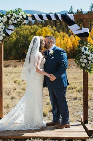 A wedding ceremony takes place under a wooden arch decorated with white flowers and striped fabric in an autumn landscape.