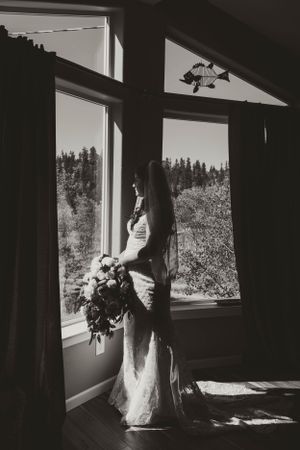 A silhouetted bride holding a bouquet stands in dramatic window light with mountain views behind her.