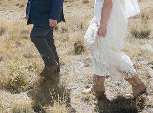 A couple in wedding attire walks through dry grass field wearing cowboy boots and formal wear.