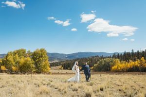 Newlyweds walking through a golden meadow with mountains and autumn trees in the background.