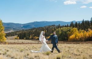 A couple walks hand in hand across a mountain meadow with fall colors and evergreen forests in the background.