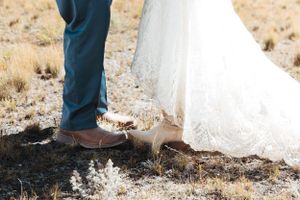 A close-up of brown dress shoes and a white wedding dress hem on dry grass.