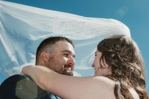 A romantic moment under a flowing white veil against a bright blue sky.